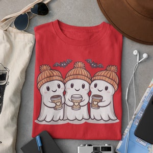 K&ouml;nnte beinhalten: Rotes T-Shirt mit einem Motiv von drei Geistern, die orangefarbene Strickm&uuml;tzen tragen und Tassen Kaffee halten. Die Geister l&auml;cheln und &uuml;ber ihnen fliegen Flederm&auml;use.