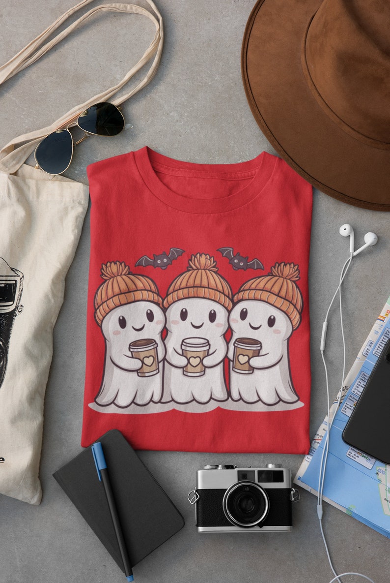 K&ouml;nnte beinhalten: Rotes T-Shirt mit einem Motiv von drei Geistern, die orangefarbene Strickm&uuml;tzen tragen und Tassen Kaffee halten. Die Geister l&auml;cheln und &uuml;ber ihnen fliegen Flederm&auml;use.