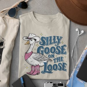 May include: A beige t-shirt with a cartoon illustration of a goose wearing a cowboy hat and boots. The text "Silly Goose on the Loose" is printed on the shirt.