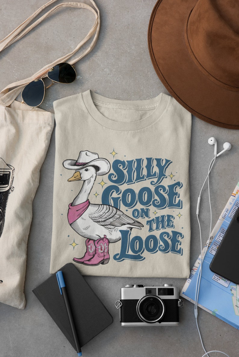 May include: A beige t-shirt with a cartoon illustration of a goose wearing a cowboy hat and boots. The text "Silly Goose on the Loose" is printed on the shirt.