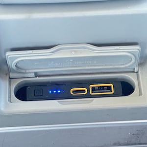 Könnte beinhalten: Eine schwarze Powerbank mit einer blauen LED-Anzeige, einem USB-C-Anschluss und einem USB-A-Anschluss. Die Powerbank befindet sich in einem grauen Fach mit einem Deckel.