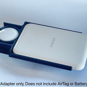 May include: Black plastic adapter for an Anker power bank. The adapter has a circular cutout for an AirTag. The text "ANKER" is visible on the power bank.