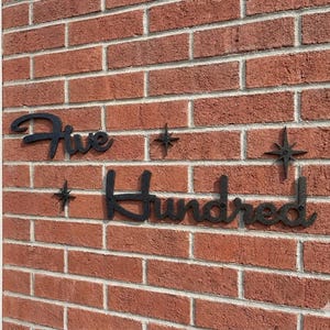 Op de afbeelding: Zwart metalen huisnummerbord met de tekst "Five Hundred" in een cursief lettertype, bevestigd aan een rode bakstenen muur. Decoratieve steraccenten zijn rond de cijfers geplaatst.