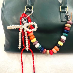 May include: A green leather handbag with a colorful beaded keychain and a red and white rope keychain. The beaded keychain features a variety of colors and textures, including white, yellow, orange, red, blue, green, and brown. The rope keychain has a heart-shaped charm.
