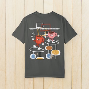 May include: A dark gray t-shirt with a retro-style graphic of two cartoon coffee cups with legs and arms. The cups are on platforms and the text "Wanna Get Breakfast?" is above them.