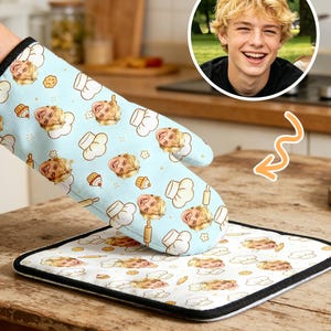 May include: A light blue oven mitt and pot holder set featuring a repeating pattern of a person's face, chef hats, and baking tools. The mitt is held up, and the pot holder is on a wooden table. The set has black trim.