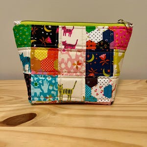 May include: A patchwork fabric zippered pouch with a variety of colorful patterns, including cats, flowers, and geometric shapes. The pouch has a lime green zipper and a gold-colored zipper pull. The pouch is sitting on a wooden surface.