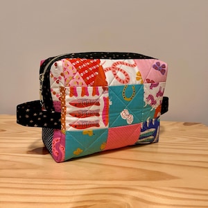 May include: A patchwork cosmetic bag with a black zipper and handle. The bag features a variety of colorful fabric squares with fish, butterflies, and horseshoe designs. The handle is black with white stars.