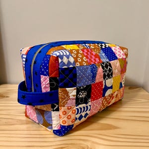 May include: A patchwork fabric zippered pouch with a blue zipper and handle. The pouch is made of various colorful fabric squares, including floral, star, and geometric patterns. The handle is blue with black star accents.