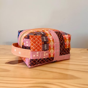 May include: A patchwork fabric cosmetic bag with a zipper closure and a handle. The bag features a variety of colorful geometric patterns in shades of pink, orange, brown, and white. The handle is a light peach color with white star accents.