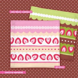 Pode incluir: Dois porta-copos quadrados de tecido medindo 10x10cm, com design inspirado em bolos. Um tem listras horizontais rosas, brancas e pêssego com morangos, enquanto o outro apresenta camadas verdes, brancas e marrons com morangos.