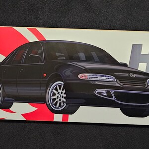 May include: A white skateboard deck with a red and white lion logo on the left side. A black Holden Commodore sedan is featured in the center of the deck. The right side of the deck has the text "HSV" in silver and a red and white lion logo in a circle.