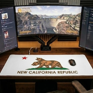 May include: A computer desk setup featuring a curved monitor displaying a video game. A white mousepad with the New California Republic flag design is in front of the monitor. A keyboard and mouse are also visible.