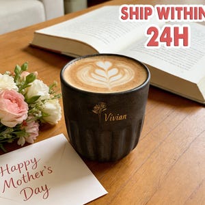 May include: A dark gray ceramic coffee cup with a latte and heart-shaped foam art, personalized with the name "Vivian". Beside it is a bouquet of pink and white flowers and a card that says "Happy Mother's Day". An open book is in the background.