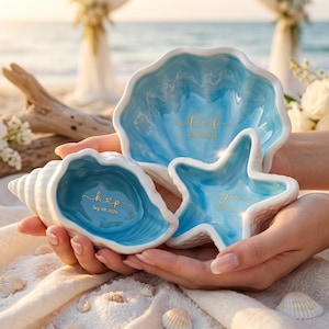 May include: Three ceramic dishes in the shape of a seashell, clam shell, and starfish. The dishes are white with a blue interior. The seashell dish has the initials "K & P" and the date "Sep 08 2026". The clam shell dish has the initials "T & F" and the date "10-10-2026". The starfish dish has the initials "Joe" and the date "08-08-2026".