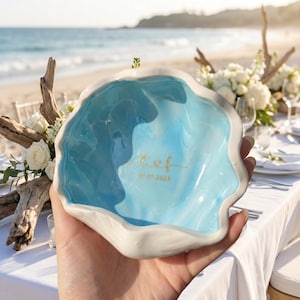 Op de afbeelding: Een blauw-witte keramische schelpvormige kom met gouden letters en de datum 07.07.2026. De kom heeft een golvende rand en wordt vastgehouden voor een strandscène met witte bloemen en drijfhout.