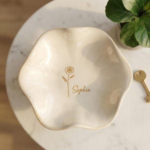May include: Cream-coloured, petal-shaped ceramic dish with a gold rose illustration and the name "Sophia" in gold script. The dish is on a marble surface, with a small plant and a gold key visible in the background.