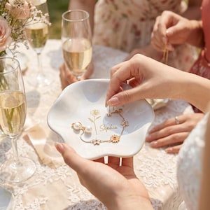 May include: A white ceramic trinket dish with a scalloped edge, featuring a gold-colored line drawing of a rose and the name Sophia printed in gold script. A delicate gold-toned chain bracelet with small flower charms and a pearl accent rests inside.
