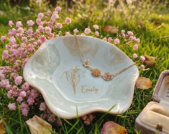 May include: A light green, flower-shaped ceramic trinket dish featuring a gold-colored line drawing of a lily of the valley and the name Emily in script. A delicate gold-tone chain bracelet with three small floral charms rests inside the dish.