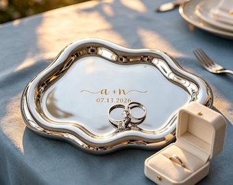 May include: A silver-colored, cloud-shaped tray with engraved text "a + n" and the date "07.13.2026". Two rings are on the tray, and a ring box with a ring inside is next to it. The tray is on a blue surface.