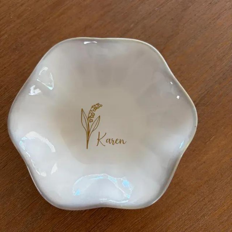 May include: A white, scalloped, flower-shaped ceramic trinket dish featuring a gold-colored illustration of a lily of the valley flower and the name Karen printed in a cursive script in the center of the bowl.