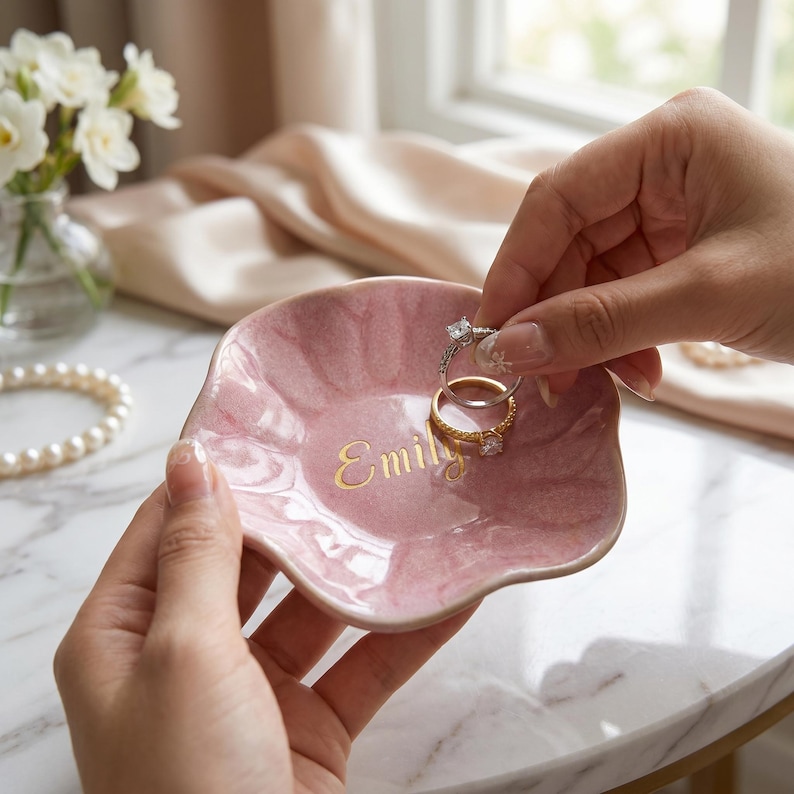 May include: A small, scalloped, pink ceramic trinket dish with a glossy finish and the name Emily printed in gold lettering. The dish is held in place while two metal rings are being placed inside the center of the bowl.