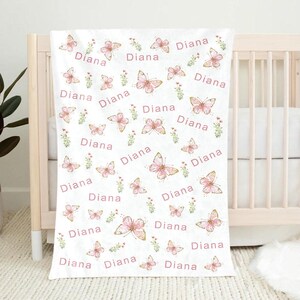 May include: White baby blanket with a repeating pattern of pink butterflies, small green floral accents, and the name "Diana" in pink script. The blanket is draped over a crib.