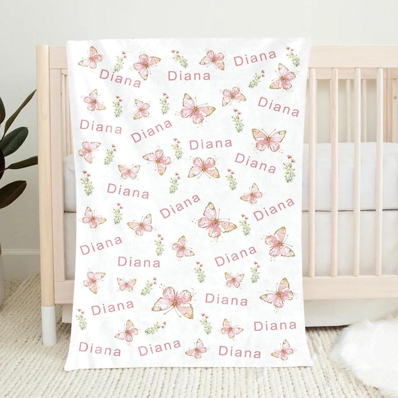 May include: White baby blanket with a repeating pattern of pink butterflies, small green floral accents, and the name "Diana" in pink script. The blanket is draped over a crib.