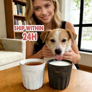 May include: Two ceramic mugs, one white and one dark gray, sit on a wooden surface. The white mug contains tea and has the name "Isaiah" and a dog silhouette. The dark gray mug has milk and the name "Lenoir" with a dog silhouette.