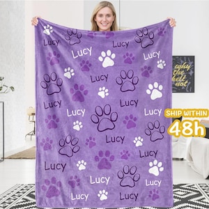 Puede incluir: Una manta de forro polar morada con un patrón de huellas de patas y el nombre "Lucy" repetido en todo. La manta es suave y acogedora, perfecta para acurrucarse en el sofá.