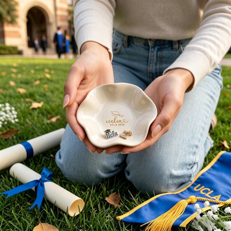 Puede incluir: Un cuenco crema con borde festoneado con letras doradas y un dise&ntilde;o de birrete. El cuenco contiene un pin de UCLA y un par de gemelos. El texto en el cuenco dice "Cecilia UCLA 2026".