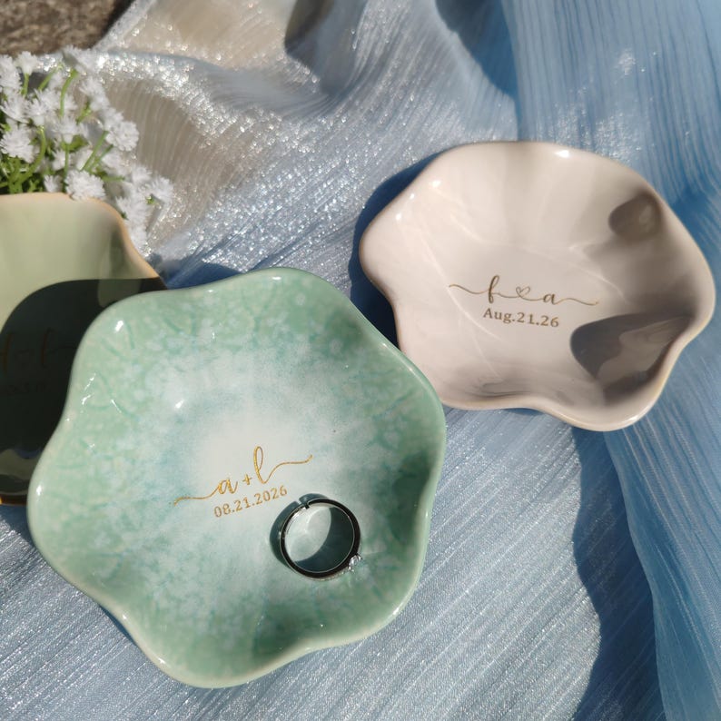 May include: Two scalloped ceramic ring dishes with gold lettering. The green dish features the text a + l 08.21.2026 and a silver ring. The beige dish features the text f + a Aug.21.26. Both dishes have a glossy, mottled finish.