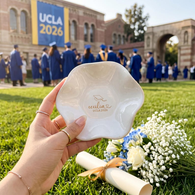 Puede incluir: Un plato de cer&aacute;mica blanco, con borde festoneado, con el texto "Cecilia UCLA 2026" en escritura dorada. El plato se sostiene frente a una ceremonia de graduaci&oacute;n con una pancarta de UCLA al fondo. Tambi&eacute;n se ve un ramo de flores y un diploma.