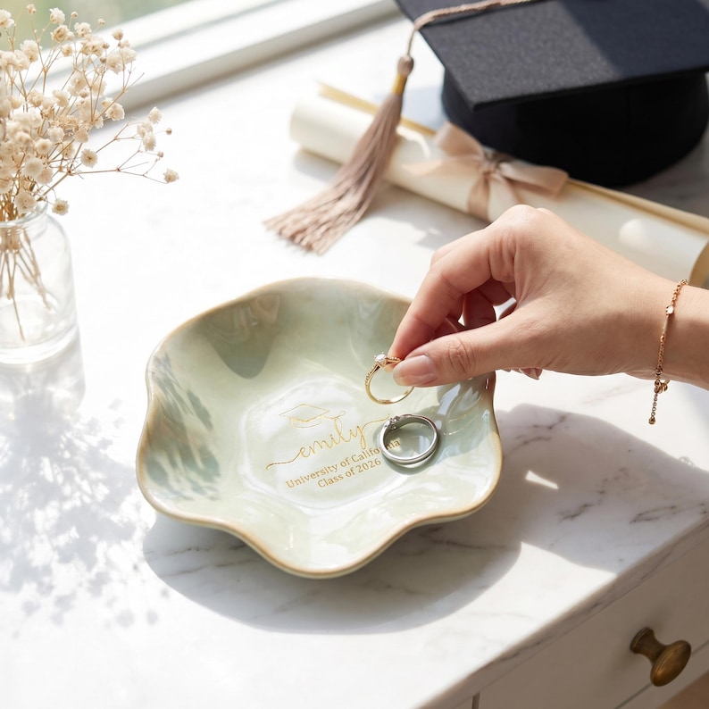 Puede incluir: Un plato decorativo verde claro con borde ondulado, que contiene dos anillos. El plato est&aacute; personalizado con el nombre "Emily" y "University of Califa Class of 2026" en letras doradas. Un birrete y un diploma est&aacute;n en el fondo.