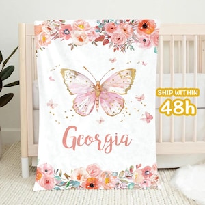 May include: A white baby blanket featuring a large pink butterfly with gold accents and the name "Georgia" in pink script. The blanket is decorated with floral arrangements in pink, peach, and coral.