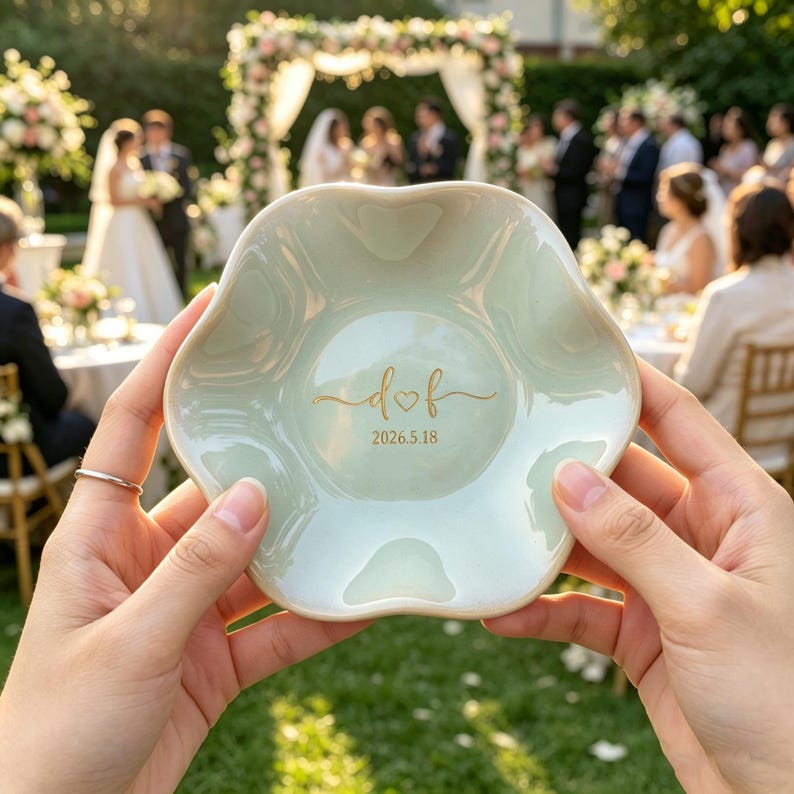 May include: A pale green, flower-shaped ceramic ring dish featuring gold lettering in the center that reads "d ♡ f" above the date "2026.5.18". The dish has a smooth, glossy finish and scalloped edges, held up against a blurred outdoor setting.