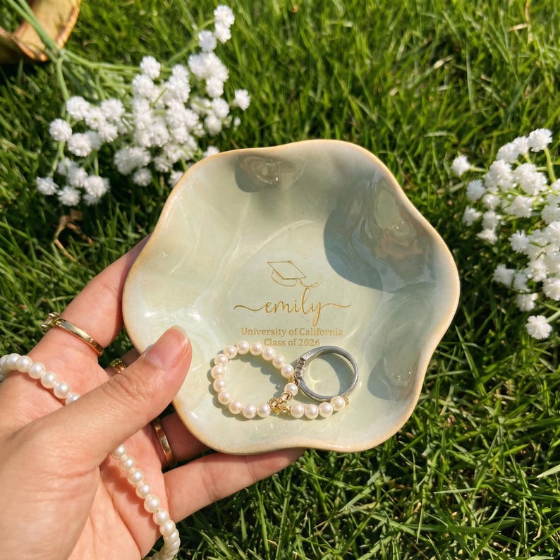 Puede incluir: Un plato de cer&aacute;mica verde claro con un borde ondulado, que contiene una pulsera de perlas y un anillo de plata. El plato est&aacute; personalizado con el nombre "emily", un gorro de graduaci&oacute;n y "University of California Class of 2026".