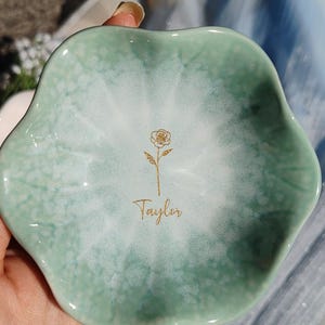 May include: A scalloped, flower-shaped ceramic trinket dish with a glossy finish. The dish features a gradient color scheme, transitioning from a soft sage green at the edges to a white center. A gold line-art rose and the name Taylor are centered.