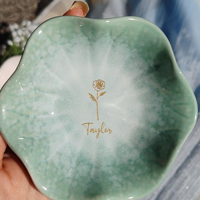 May include: A scalloped, flower-shaped ceramic trinket dish with a glossy finish. The dish features a gradient color scheme, transitioning from a soft sage green at the edges to a white center. A gold line-art rose and the name Taylor are centered.