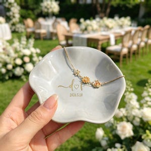 May include: A light gray, flower-shaped ceramic trinket dish featuring gold-colored text that reads d heart f and 2026.5.18. A delicate gold-tone chain necklace with three small floral charms rests inside the dish.