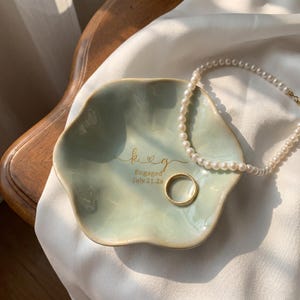 May include: A pale green, flower-shaped ceramic trinket dish featuring the gold-colored text k & g, Engaged, and July 21.26. A simple gold band and a white pearl necklace are resting inside the dish on its smooth, glossy surface.