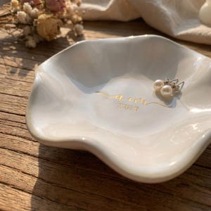 May include: A white ceramic ring dish with a wavy edge. The dish has gold lettering that reads "a + m" and the date "2020.9.13". Two pearl earrings rest inside the dish. The dish is on a wooden surface.