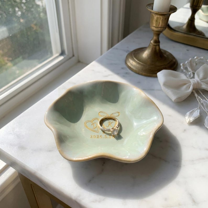 May include: A pale green, scalloped ceramic ring dish featuring gold-colored text and a heart graphic with the letters D and K, along with the date 2024.5.18. A gold-toned metal ring with a clear stone sits in the center of the dish.