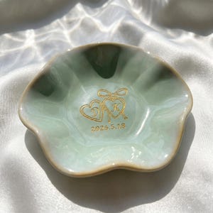 May include: A pale green ceramic trinket dish with a scalloped edge. The center features a gold-colored graphic of two connected hearts with the letters D and K inside, topped with a bow, and the date 2026.5.18 printed in gold below the hearts.