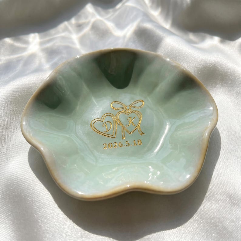 May include: A pale green ceramic trinket dish with a scalloped edge. The center features a gold-colored graphic of two connected hearts with the letters D and K inside, topped with a bow, and the date 2026.5.18 printed in gold below the hearts.