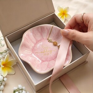 Puede incluir: Un cuenco de cer&aacute;mica rosa con un collar dorado dentro de una caja de regalo. El cuenco tiene el nombre "ashley" y el a&ntilde;o "2026" en letras doradas. El collar tiene colgantes de flores y estrellas. Se ve una cinta rosa.