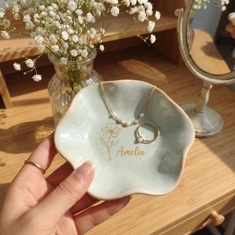 May include: A light blue, flower-shaped ceramic trinket dish featuring a gold-colored line drawing of a flower and the name Amelia printed in gold script. The dish holds a delicate gold chain necklace with small pearls and a gold ring with a pearl.