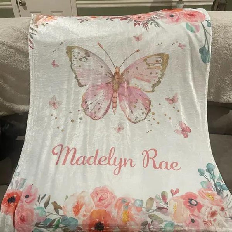 May include: A white blanket featuring a large pink butterfly with gold accents and the name "Madelyn Rae" in pink script. The blanket is decorated with floral and butterfly designs in shades of pink and peach.