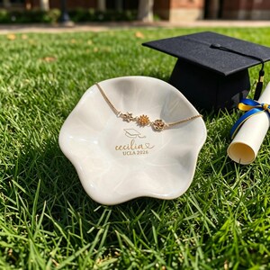 Puede incluir: Un plato de cer&aacute;mica blanco con una pulsera floral dorada, un birrete y un diploma sobre hierba verde. El plato est&aacute; inscrito con "Cecilia UCLA 2026". La pulsera presenta tres dijes de flores. El diploma est&aacute; atado con cintas azules y amarillas.