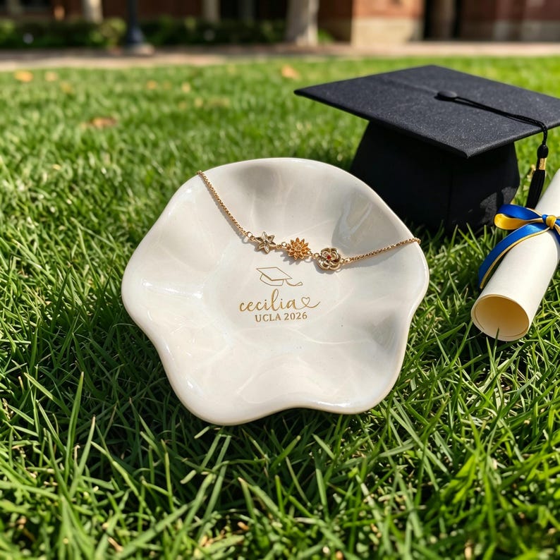 Puede incluir: Un plato de cer&aacute;mica blanco con una pulsera floral dorada, un birrete y un diploma sobre hierba verde. El plato est&aacute; inscrito con "Cecilia UCLA 2026". La pulsera presenta tres dijes de flores. El diploma est&aacute; atado con cintas azules y amarillas.